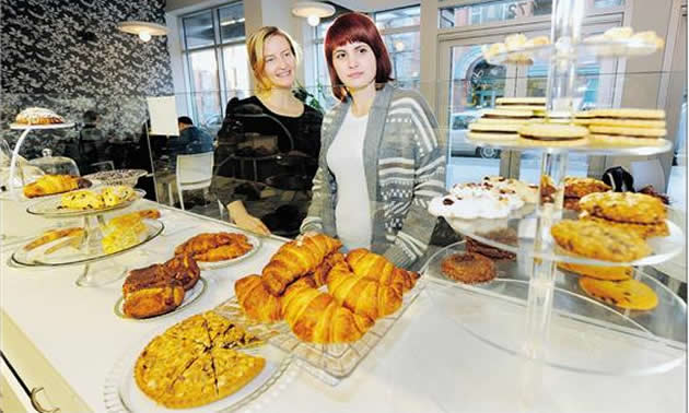 Two women stand behind a white counter covered in delicious-looking golden pastries.