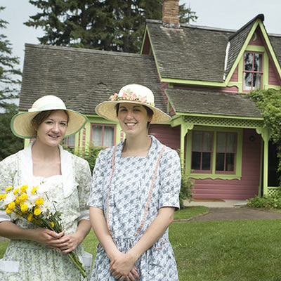 Hisotric house, with two ladies dressed in period costume in the foreground, one of them holding a bouquet of yellow daisies. 