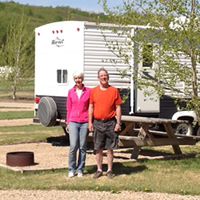 A couple stand in an attractive campsite with a small tree, a fire ring, and an RV behind them.