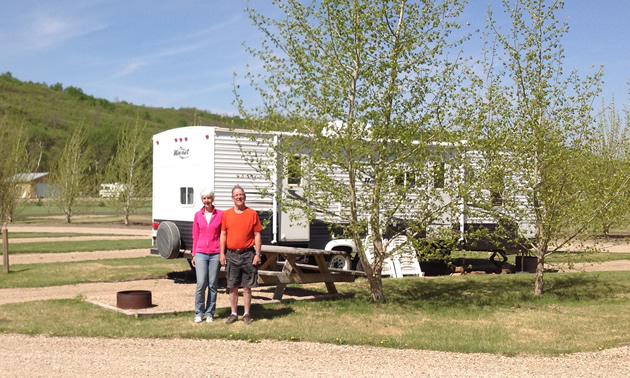 A couple stand in an attractive campsite with a small tree, a fire ring, and an RV behind them.