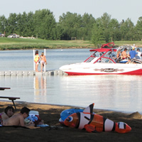 Sunbathers on a beach in the foreground, children and a power boat in the water in the mid-ground and a grassy slope in the far background