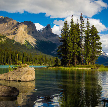 Spirit island in Maligne Lake is basking in the sun.  The waters from the island to the shore are calm and reflecting the island and mountains behind. 